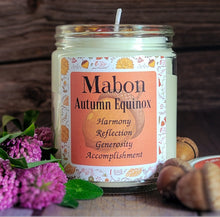 Load image into Gallery viewer, A glass jar candle labeled 'Mabon Autumn Equinox' with a white wax fill, autumn-themed label, and wick, styled on a wooden surface with purple flowers, acorns, and a small dish in the foreground.