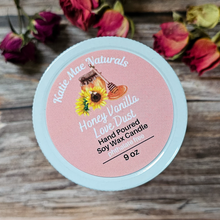 Load image into Gallery viewer, Candle container with pink label featuring 'Katie Mae Naturals' on a wooden surface with flowers.
